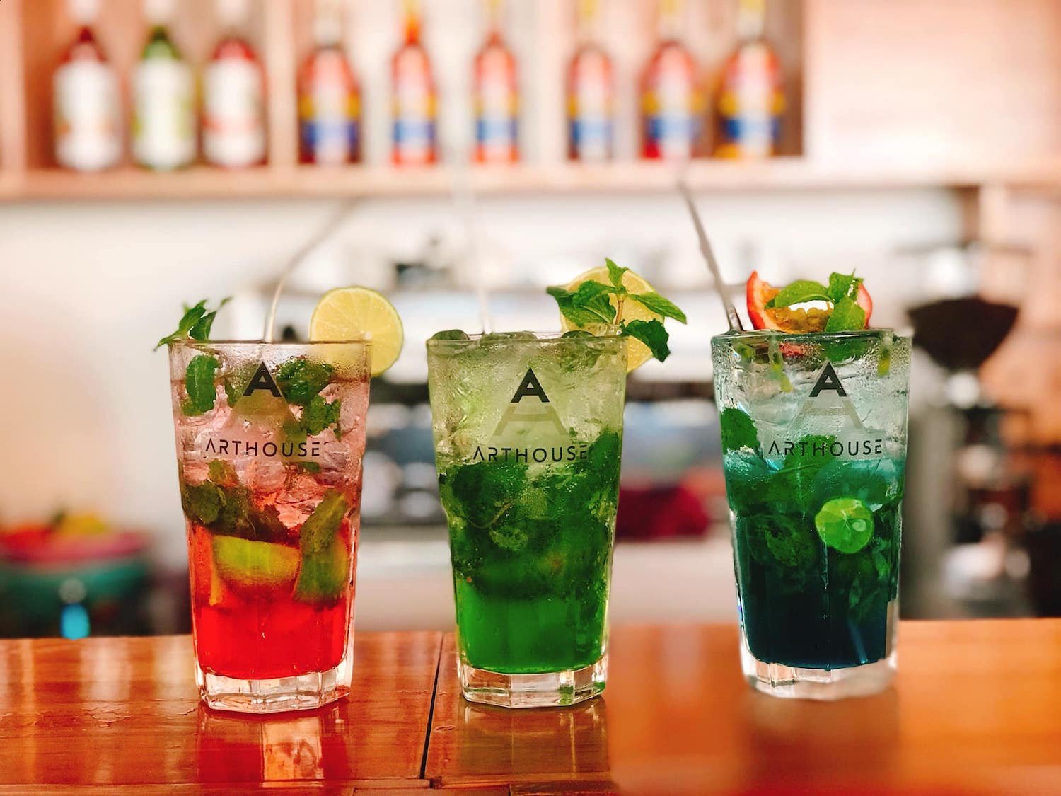 Ba ly cocktail màu sắc khác nhau với đá và trái cây, đặt trên quầy gỗ, nền là giá rượu mờ.