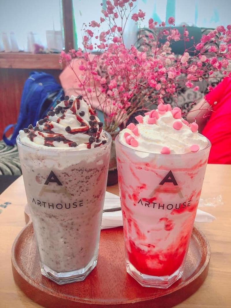 Hai ly đồ uống kem tươi trang trí với siro và socola, đặt trên bàn gỗ gần hoa khô màu hồng.