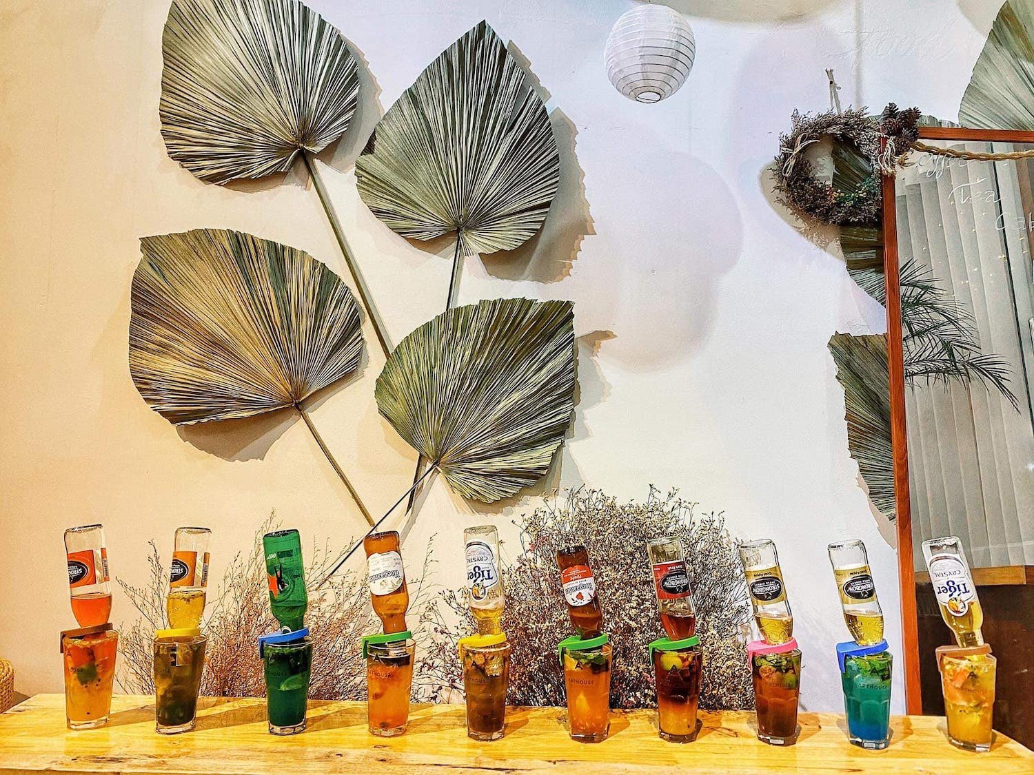 Các ly đồ uống trên bàn gỗ với chai bia úp ngược, trang trí lá khô trên tường phía sau.