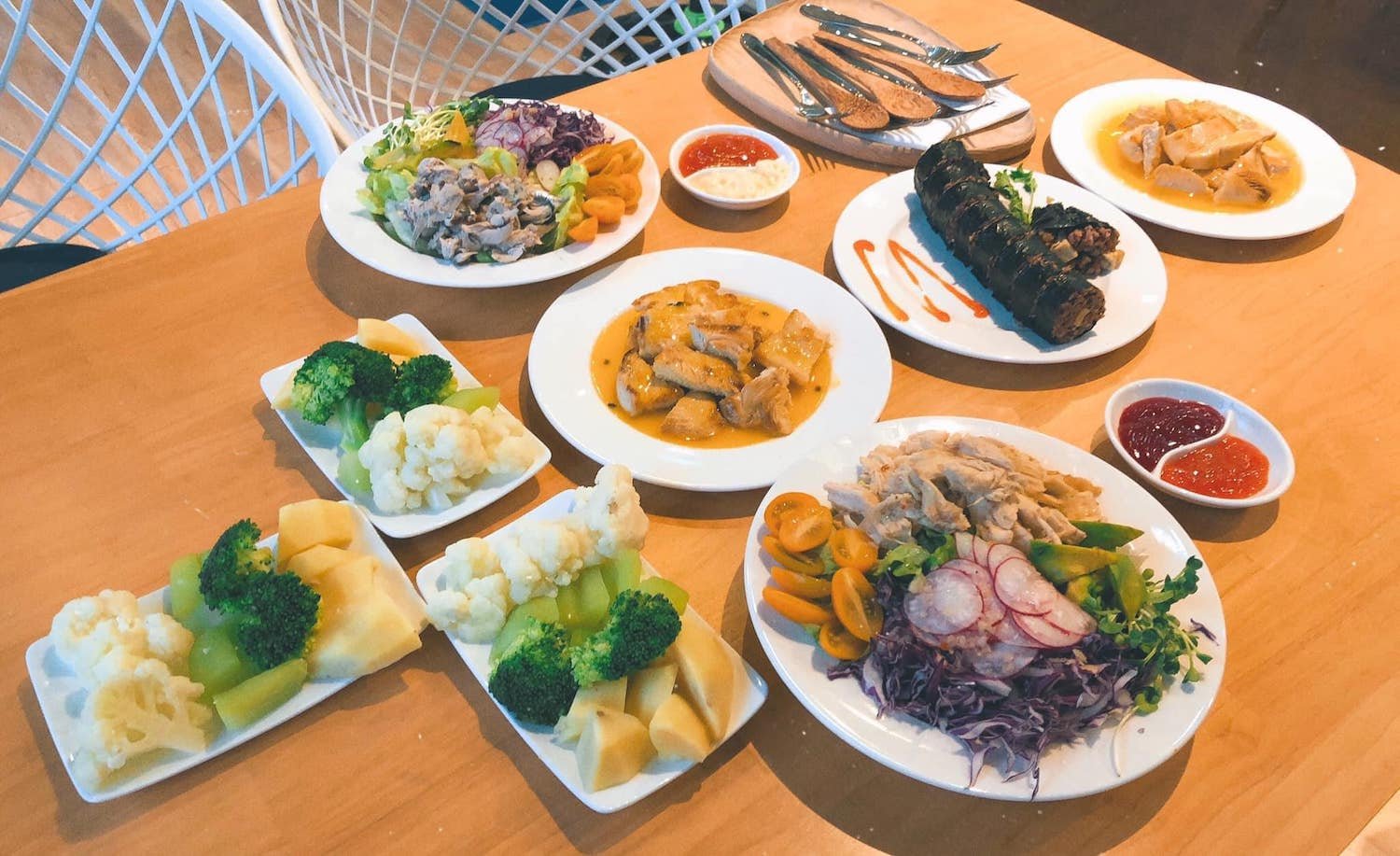 Bàn ăn với nhiều món ăn đa dạng gồm rau củ, thịt gà, cuốn lá và nước chấm trên nền gỗ sáng màu.