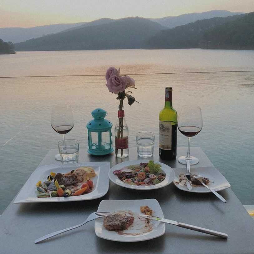 B&agrave;n ăn ngo&agrave;i trời với rượu vang, thịt b&ograve;, salad, hoa v&agrave; đ&egrave;n lồng xanh, nh&igrave;n ra hồ v&agrave; n&uacute;i l&uacute;c ho&agrave;ng h&ocirc;n.