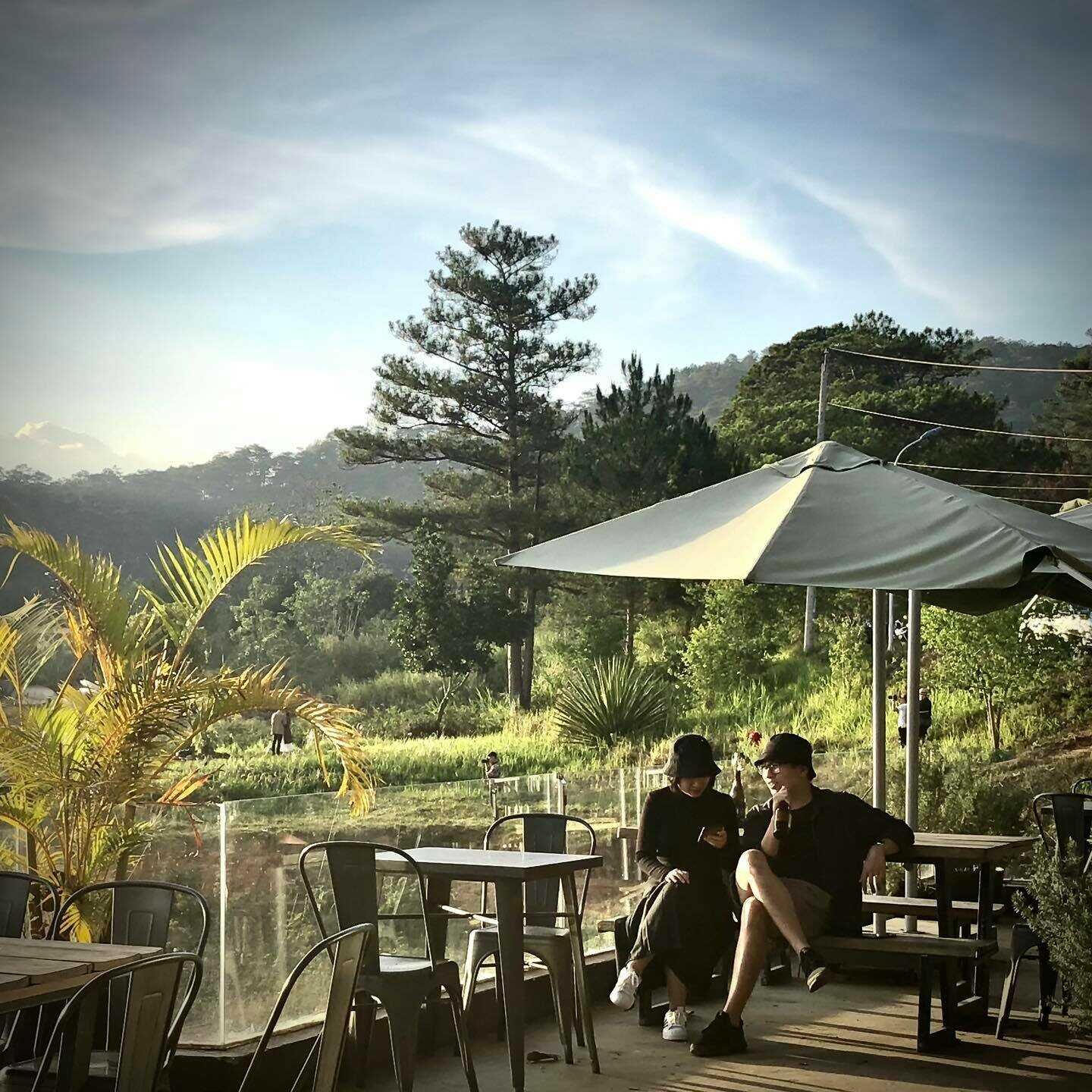 Hai người ngồi dưới &ocirc; che nắng tại qu&aacute;n c&agrave; ph&ecirc; ngo&agrave;i trời, xung quanh l&agrave; c&acirc;y xanh v&agrave; n&uacute;i đồi.