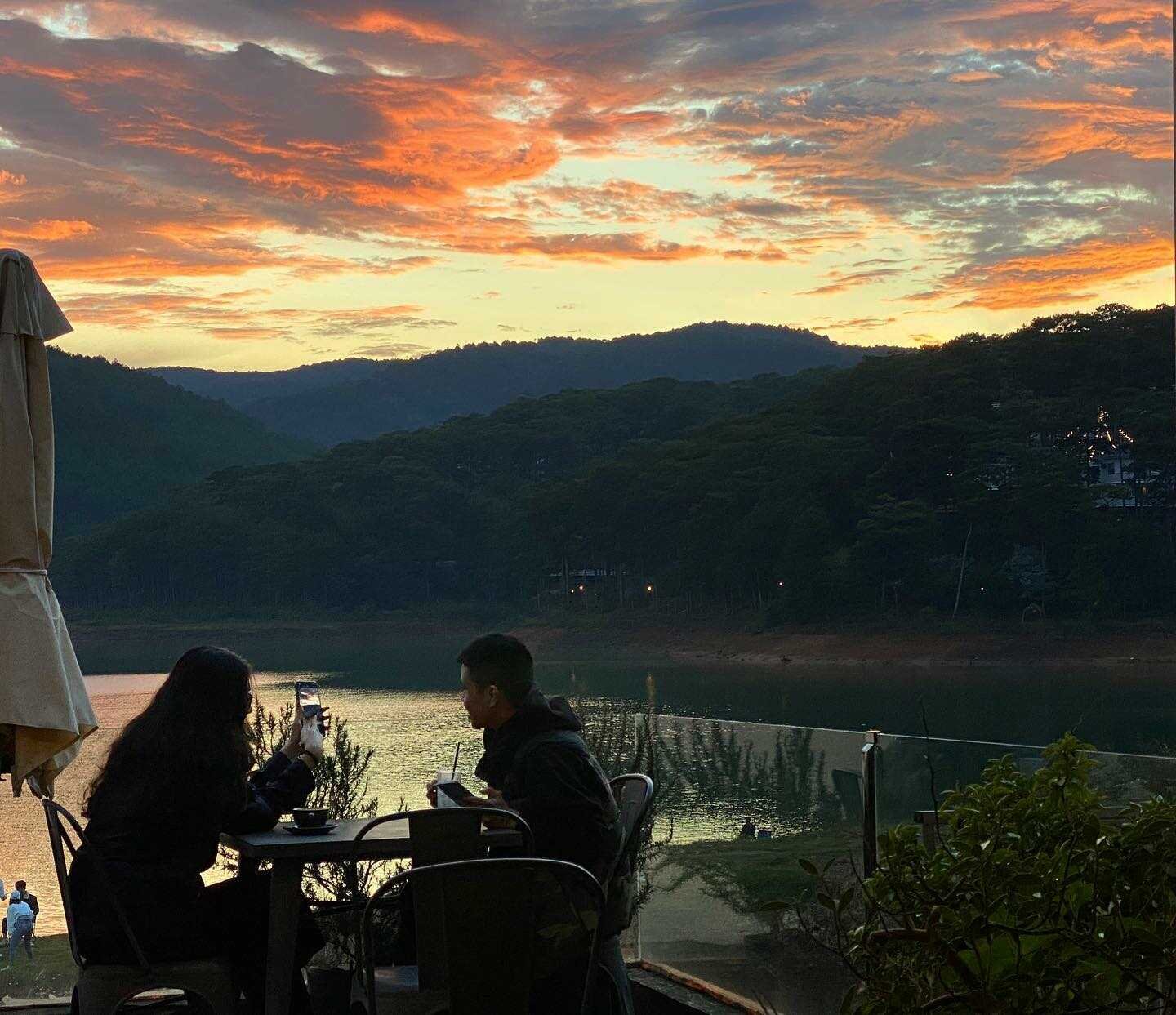 Hai người ngồi tại b&agrave;n ngo&agrave;i trời, nh&igrave;n ra hồ nước l&uacute;c ho&agrave;ng h&ocirc;n, với bầu trời rực rỡ sắc cam v&agrave; hồng.