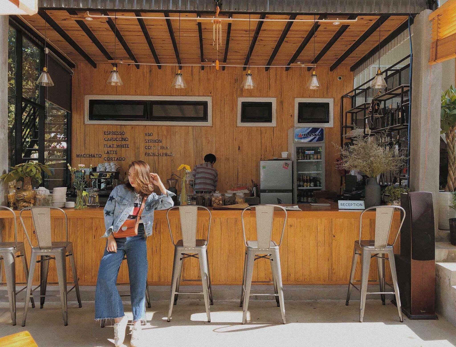 Qu&aacute;n c&agrave; ph&ecirc; với quầy gỗ, ghế cao v&agrave; tấm bảng menu, c&oacute; người đứng ph&iacute;a trước v&agrave; ph&iacute;a sau quầy.