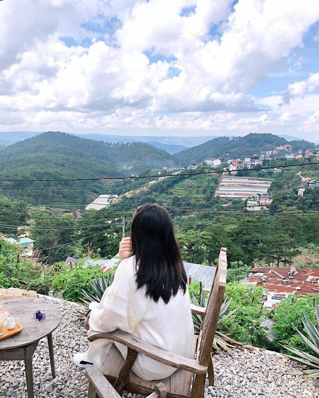 Người ngồi tr&ecirc;n ghế gỗ, ngắm nh&igrave;n khung cảnh đồi n&uacute;i xanh v&agrave; bầu trời nhiều m&acirc;y từ một qu&aacute;n c&agrave; ph&ecirc; ngo&agrave;i trời.