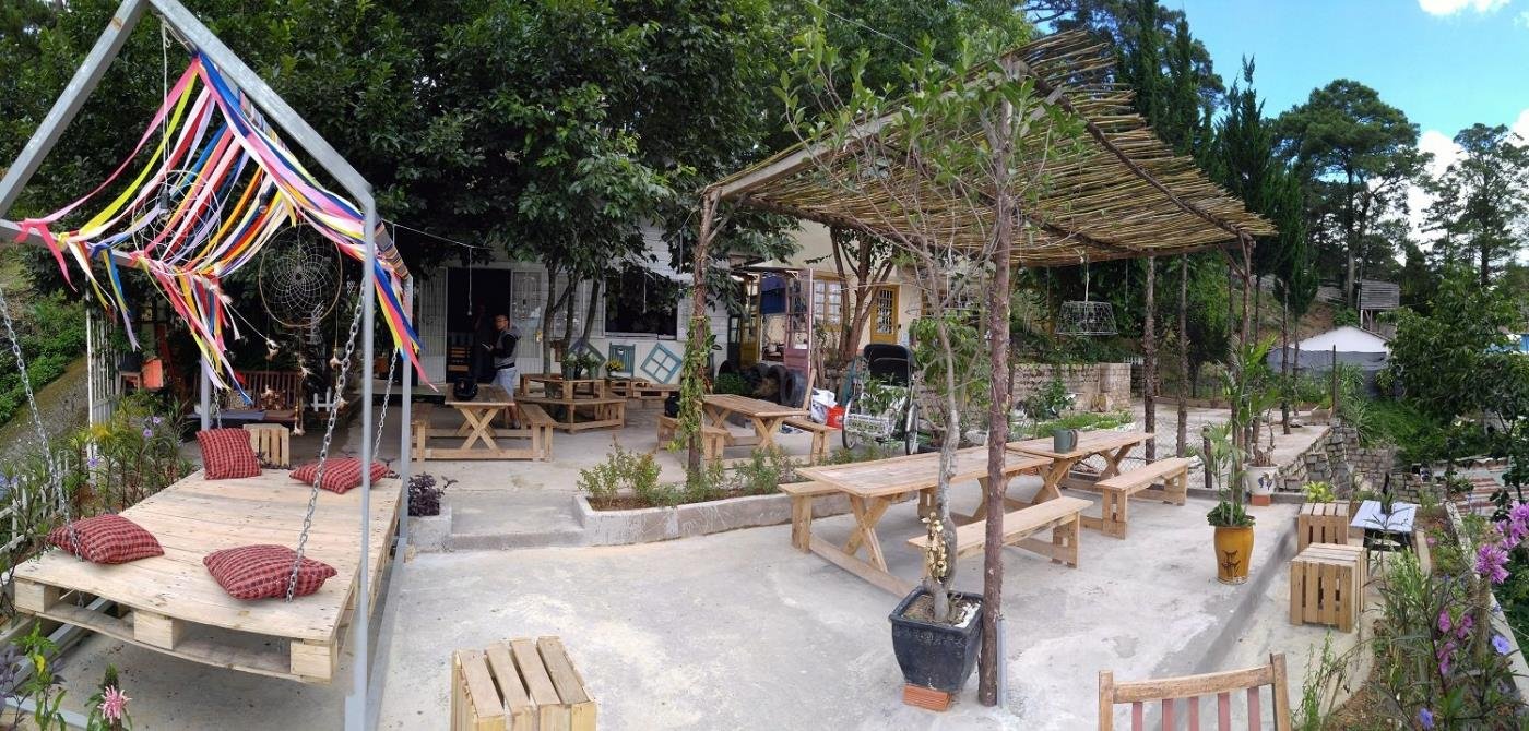 Khu vườn ngo&agrave;i trời với b&agrave;n ghế gỗ, m&aacute;i che tre, v&agrave; trang tr&iacute; d&acirc;y m&agrave;u sắc, c&acirc;y cối xung quanh xanh tươi.