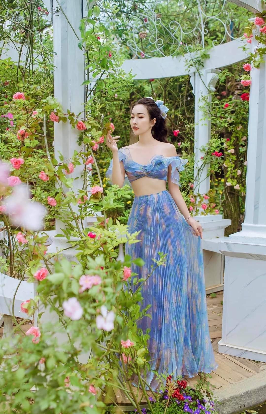 Người phụ nữ mặc v&aacute;y xanh đứng trong vườn hoa hồng, xung quanh l&agrave; cột trắng v&agrave; c&acirc;y cối xanh tươi.