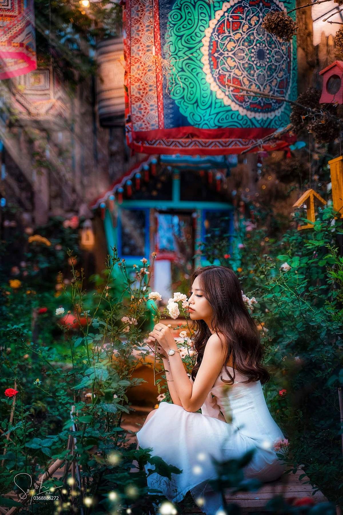 Người phụ nữ trong vườn hoa, ngồi cạnh b&ocirc;ng hồng, ph&ocirc;ng nền trang tr&iacute; bằng vải m&agrave;u sắc.