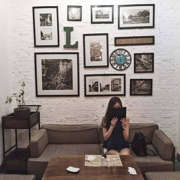 Người ngồi trên ghế sofa trong quán cà phê, cầm thực đơn. Tường trang trí khung ảnh và đồng hồ.