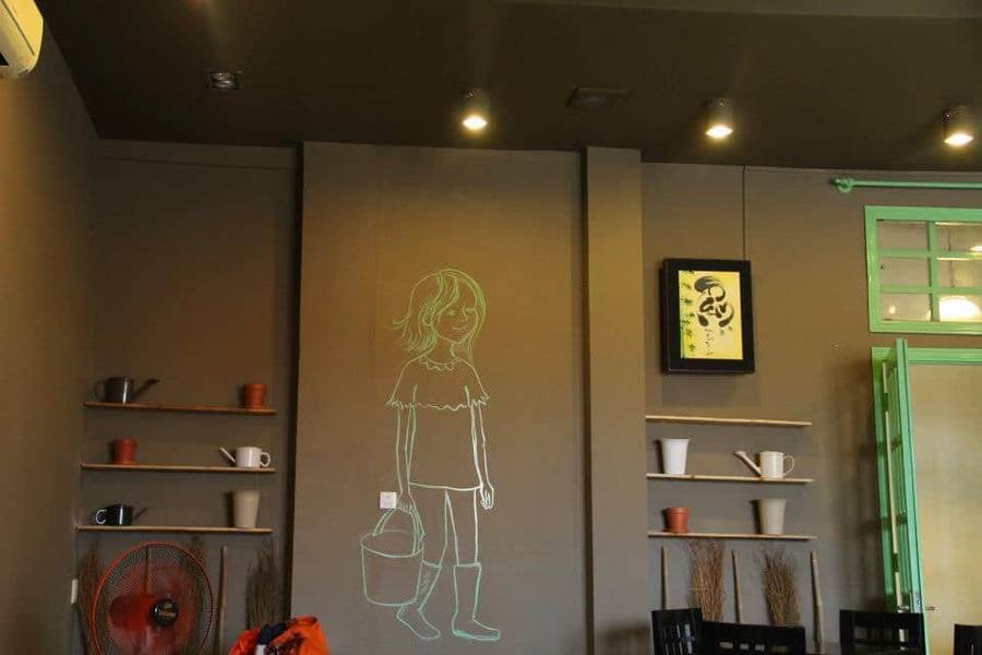 Bức tường quán cà phê với tranh vẽ cô gái cầm xô, bên cạnh là tranh treo và kệ có cốc, bình trang trí.