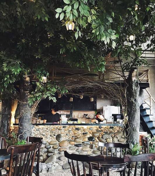 Quán cà phê với quầy bar bằng đá, cây xanh xung quanh và bàn ghế gỗ, tạo không gian gần gũi với thiên nhiên.