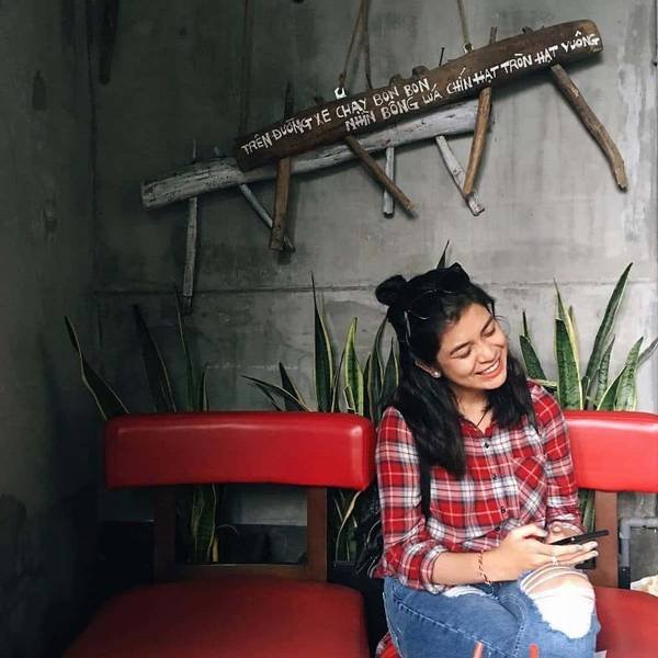 Người phụ nữ ngồi trên ghế đỏ, mặc áo sơ mi kẻ ca rô, tay cầm điện thoại, phía sau là tường xám và cây xanh.