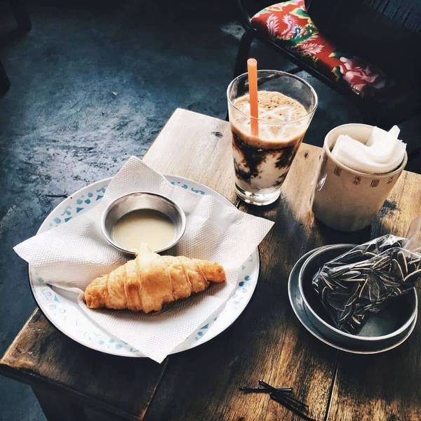 Bàn gỗ với bánh sừng bò, ly cà phê sữa đá, kèm chén sữa đặc, và túi trà trên đĩa nhỏ.