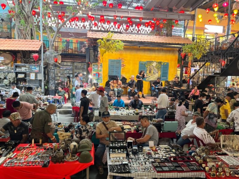 Chợ đồ cổ đ&ocirc;ng đ&uacute;c với nhiều gian h&agrave;ng b&agrave;y b&aacute;n đồng hồ, n&oacute;n v&agrave; c&aacute;c vật dụng kh&aacute;c, c&oacute; ban nhạc biểu diễn ph&iacute;a sau.