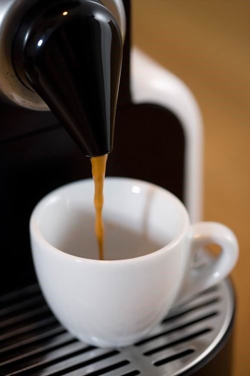 Máy pha cà phê đang rót cà phê vào tách sứ trắng trên khay kim loại.
