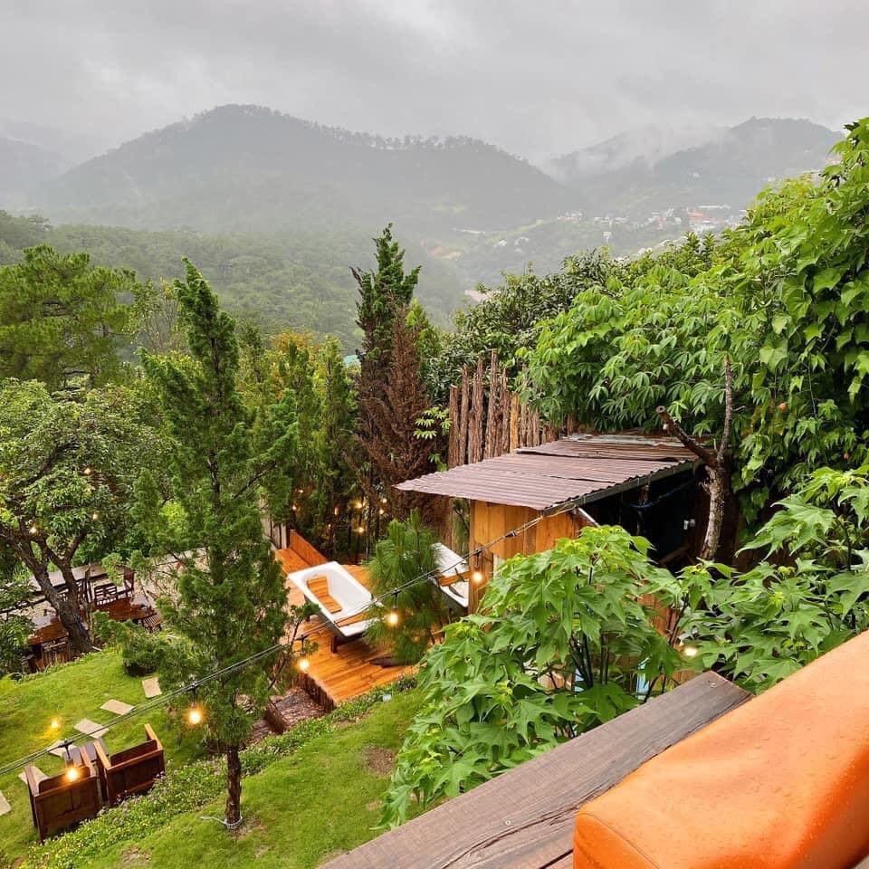 Khu vườn xanh tươi với ghế thư gi&atilde;n, nh&agrave; gỗ v&agrave; đ&egrave;n trang tr&iacute;, n&uacute;i non mờ ảo trong sương m&ugrave; ph&iacute;a xa.