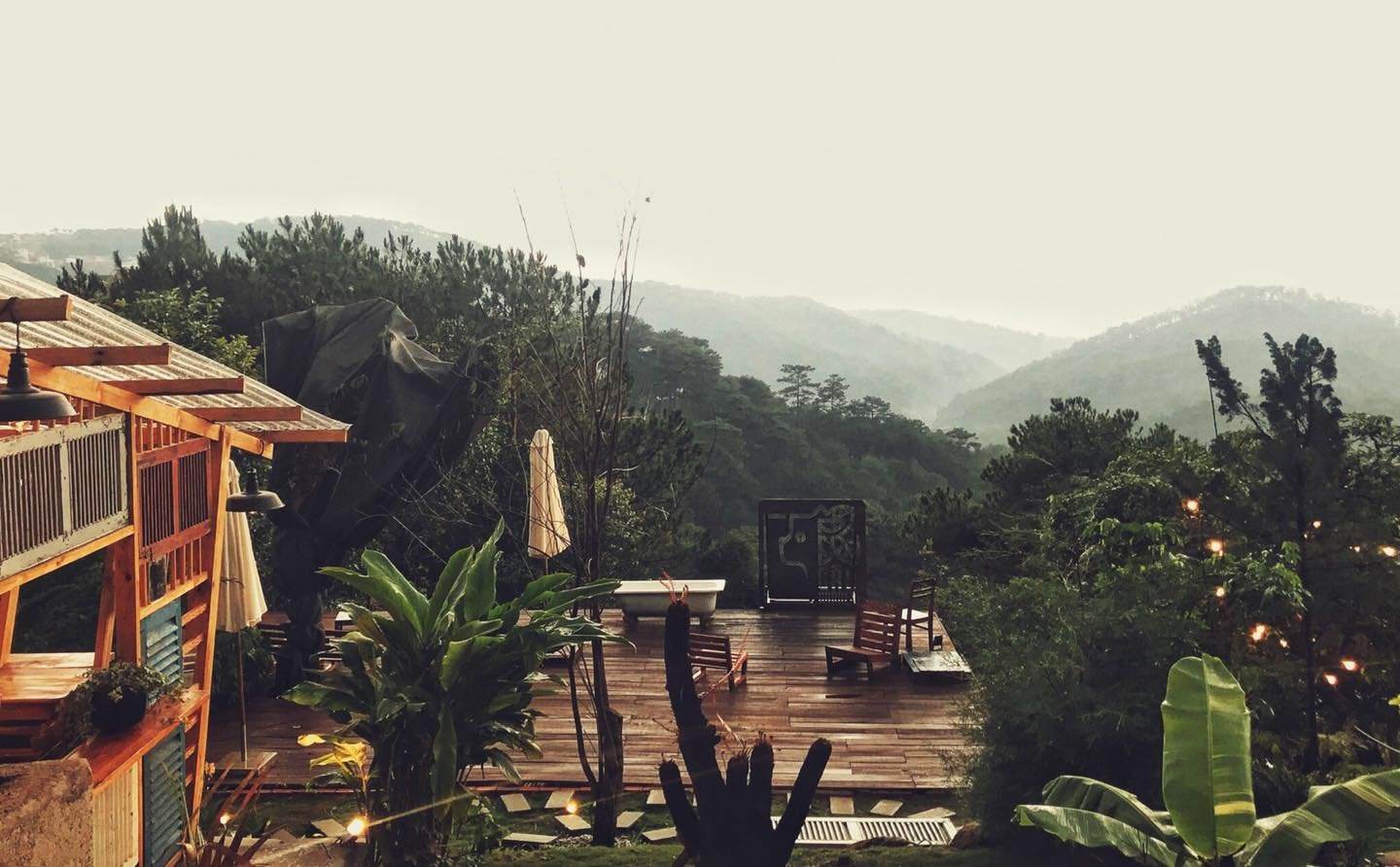 Ng&ocirc;i nh&agrave; gỗ b&ecirc;n sườn đồi, ph&iacute;a trước l&agrave; s&acirc;n gỗ, c&acirc;y xanh tươi tốt, nền l&agrave; d&atilde;y n&uacute;i phủ sương mờ.