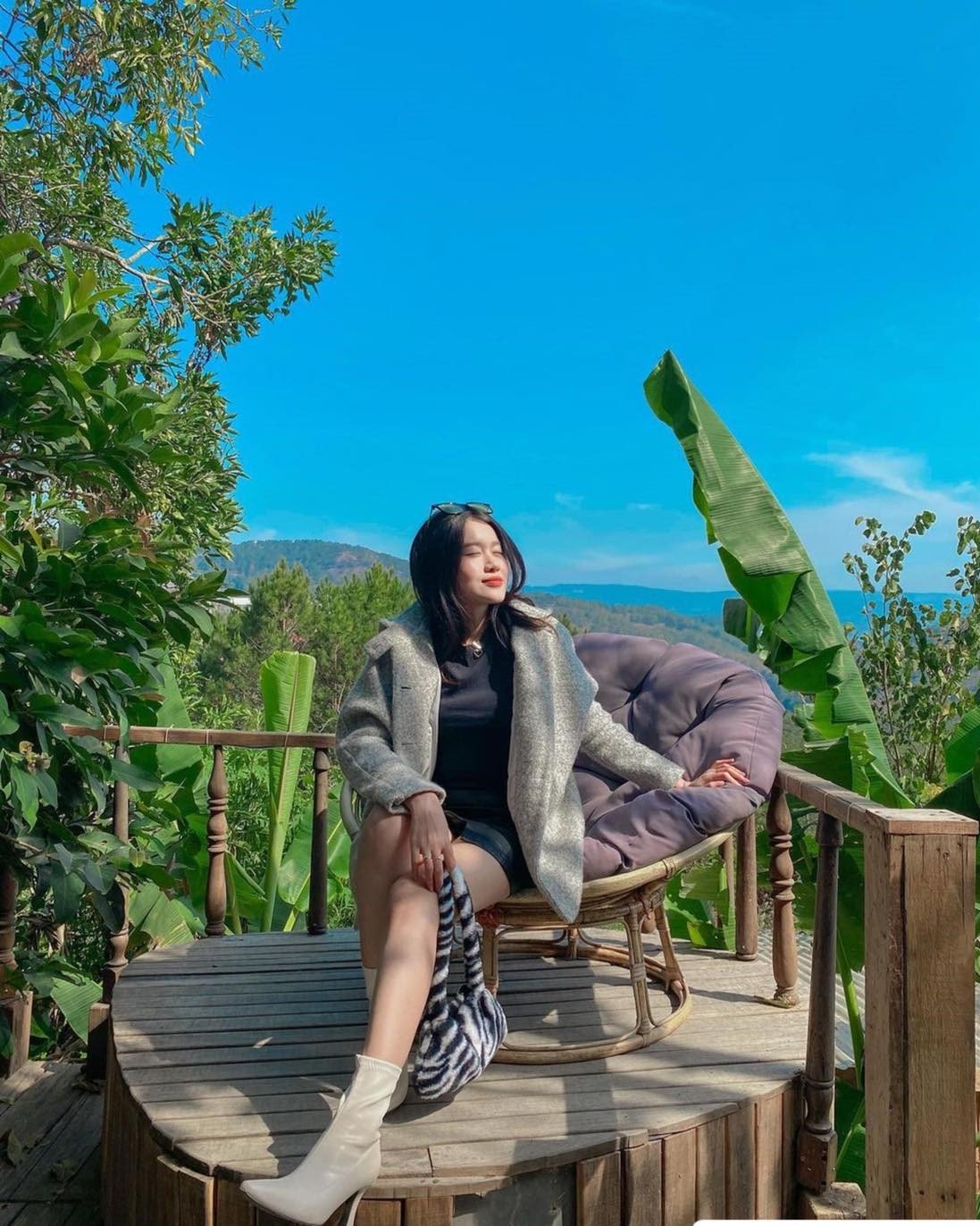 Người phụ nữ ngồi tr&ecirc;n ghế ngo&agrave;i trời, tận hưởng &aacute;nh nắng. Xung quanh l&agrave; c&acirc;y xanh v&agrave; bầu trời trong xanh.