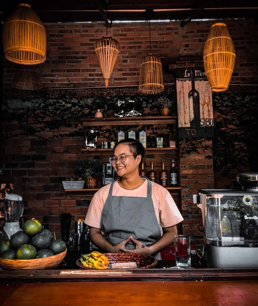 Người đứng sau quầy pha chế với giỏ tr&aacute;i c&acirc;y v&agrave; m&aacute;y pha c&agrave; ph&ecirc;, trong kh&ocirc;ng gian trang tr&iacute; ấm c&uacute;ng.