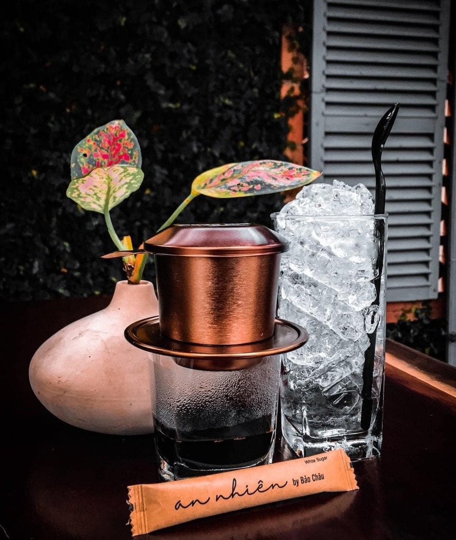 Phin c&agrave; ph&ecirc; đặt tr&ecirc;n ly nước, b&ecirc;n cạnh l&agrave; ly đ&aacute; v&agrave; b&igrave;nh hoa nhỏ với l&aacute; c&acirc;y trang tr&iacute;.