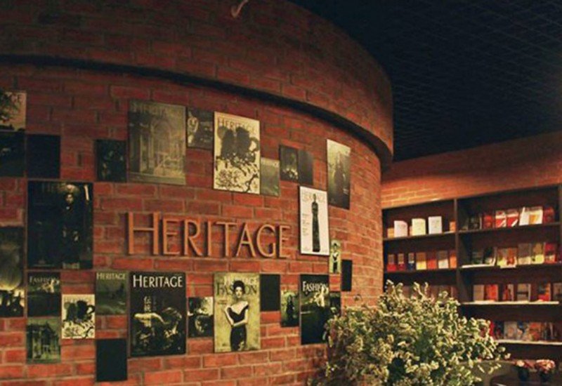 Bức tường gạch trang tr&iacute; với nhiều tạp ch&iacute; v&agrave; chữ "HERITAGE", b&ecirc;n cạnh l&agrave; kệ s&aacute;ch v&agrave; một chậu c&acirc;y xanh.