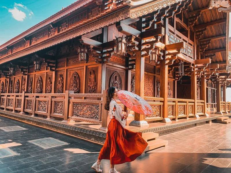 Người phụ nữ cầm &ocirc; đứng trước ng&ocirc;i ch&ugrave;a gỗ truyền thống, &aacute;nh s&aacute;ng chiếu rọi tạo n&ecirc;n khung cảnh ấm &aacute;p.