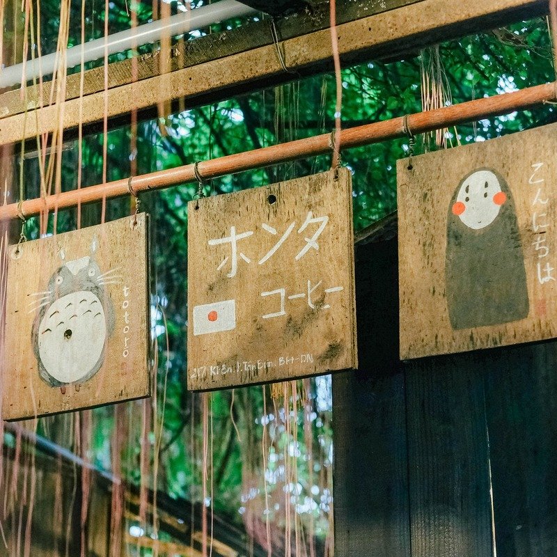 Ba biển hiệu gỗ với h&igrave;nh vẽ v&agrave; chữ Nhật, treo trong kh&ocirc;ng gian ngo&agrave;i trời c&oacute; c&acirc;y xanh.