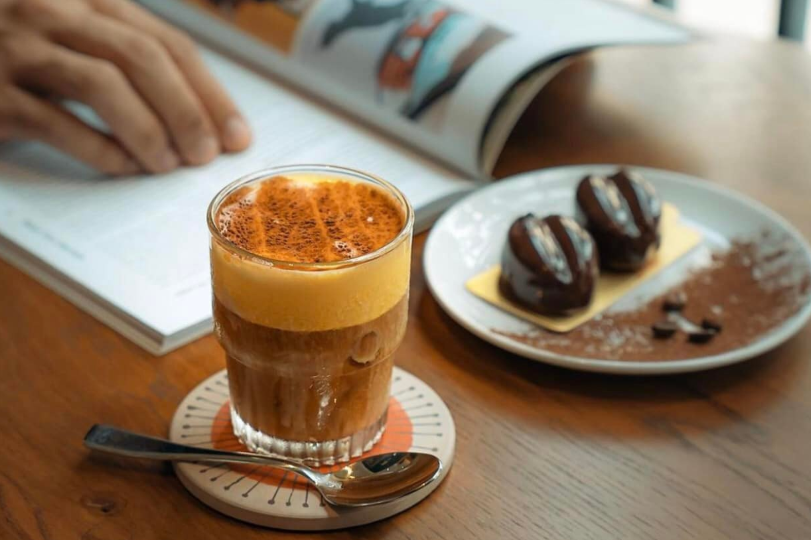 Ly cà phê trứng và đĩa bánh ngọt trên bàn gỗ, bên cạnh là tay người lật trang sách.