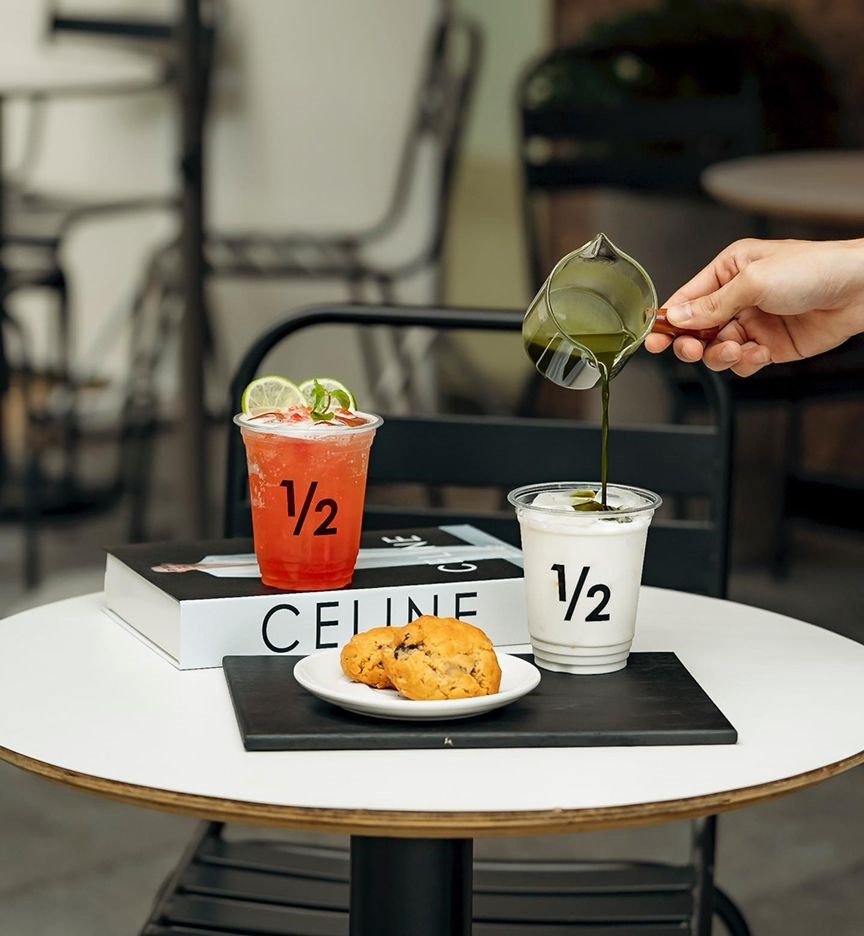 Cốc nước màu đỏ và trắng trên bàn, một người đang rót chất lỏng xanh vào cốc trắng, bên cạnh là đĩa bánh.