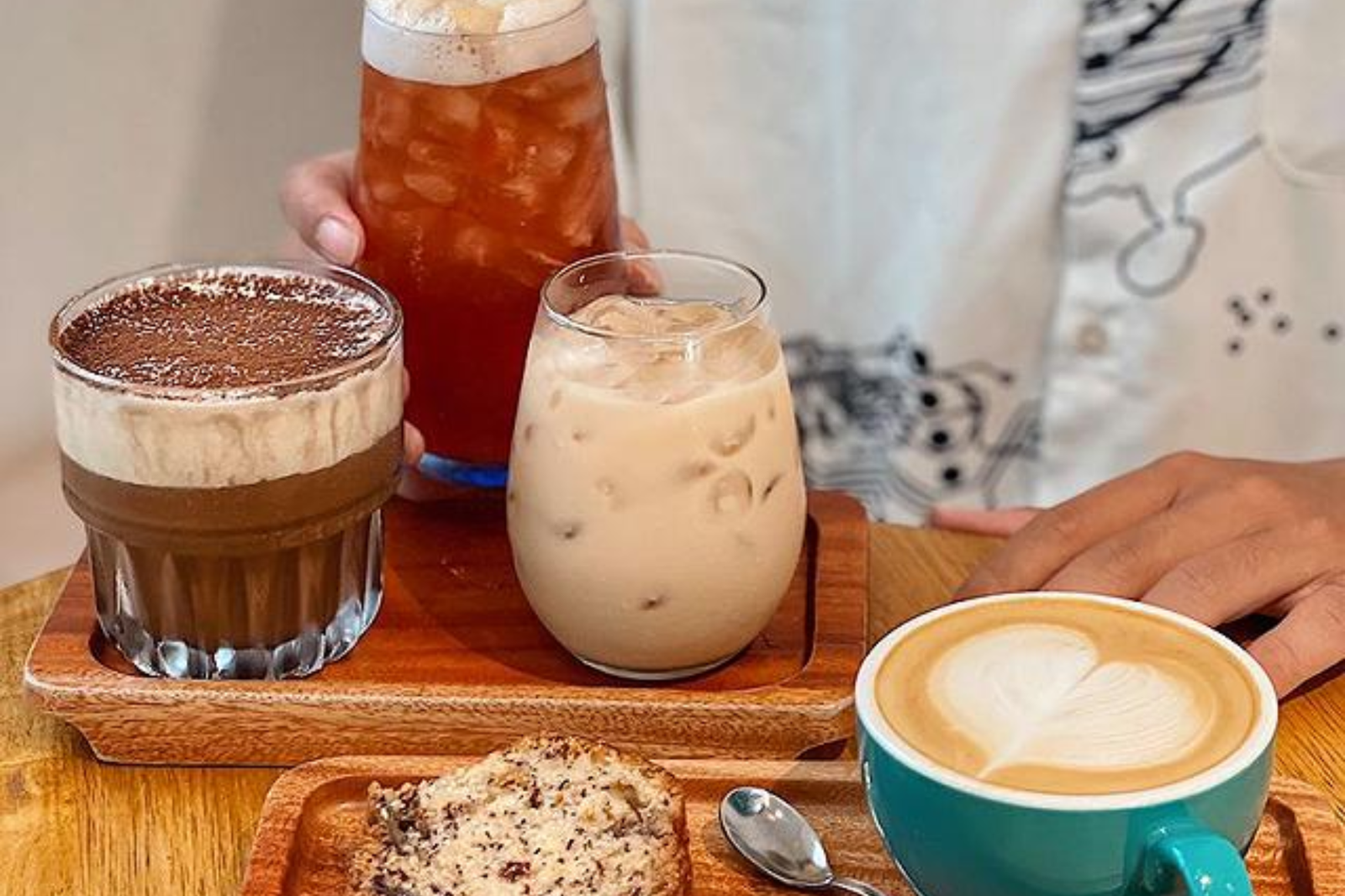 Bàn gỗ với các loại đồ uống: cà phê, trà đá, sữa đá và một chiếc bánh nhỏ, cùng một người đang cầm ly.