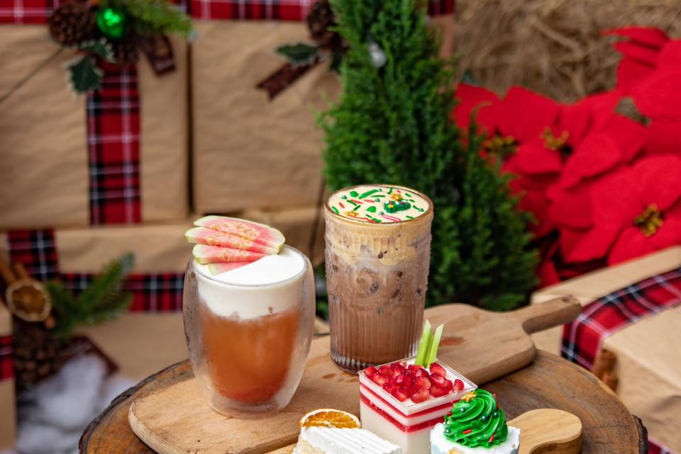 Hai ly đồ uống và ba miếng bánh trên bàn gỗ, nền có cây xanh và hoa đỏ trang trí.