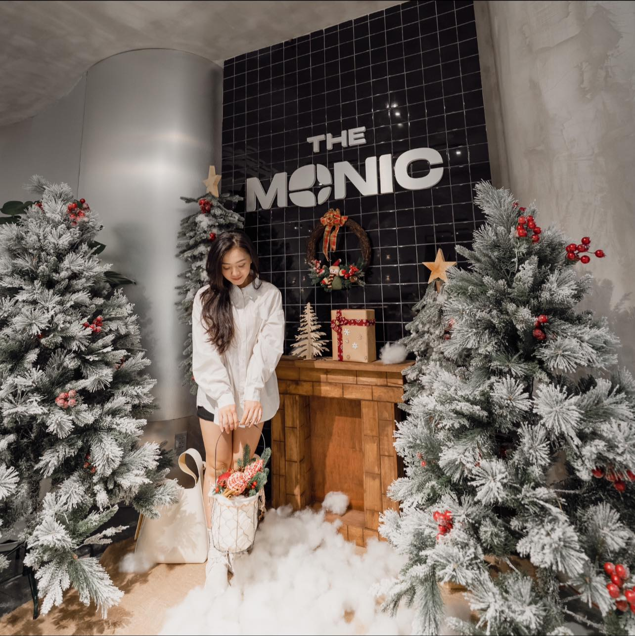 Người đứng cạnh cây thông Noel trang trí, trong không gian có nền gạch đen và lò sưởi gỗ.