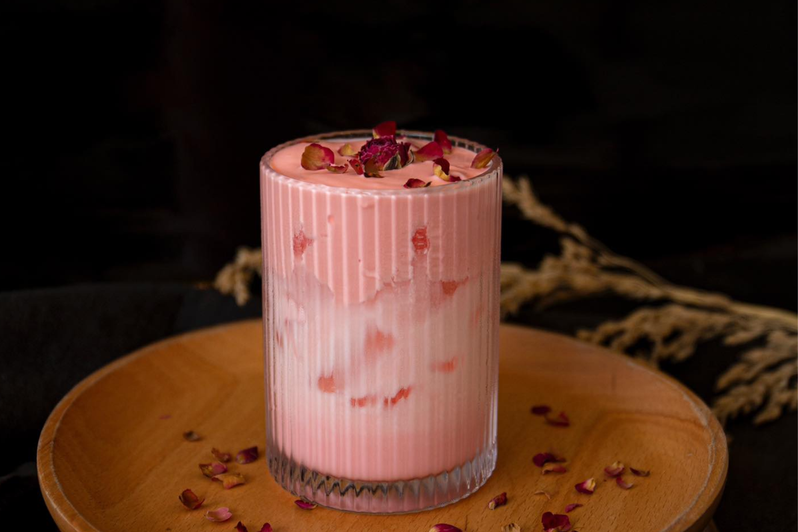Ly đồ uống màu hồng với hoa hồng khô trên mặt, đặt trên đĩa gỗ.