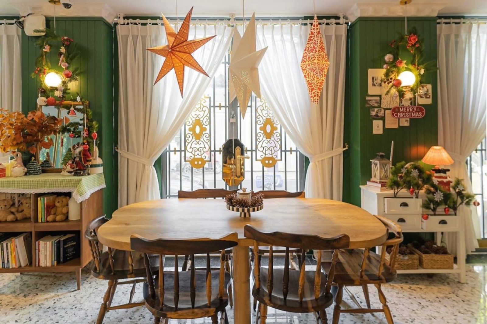 Phòng ăn trang trí Giáng Sinh với bàn gỗ, ghế, đèn hình ngôi sao, cây thông nhỏ và rèm trắng.