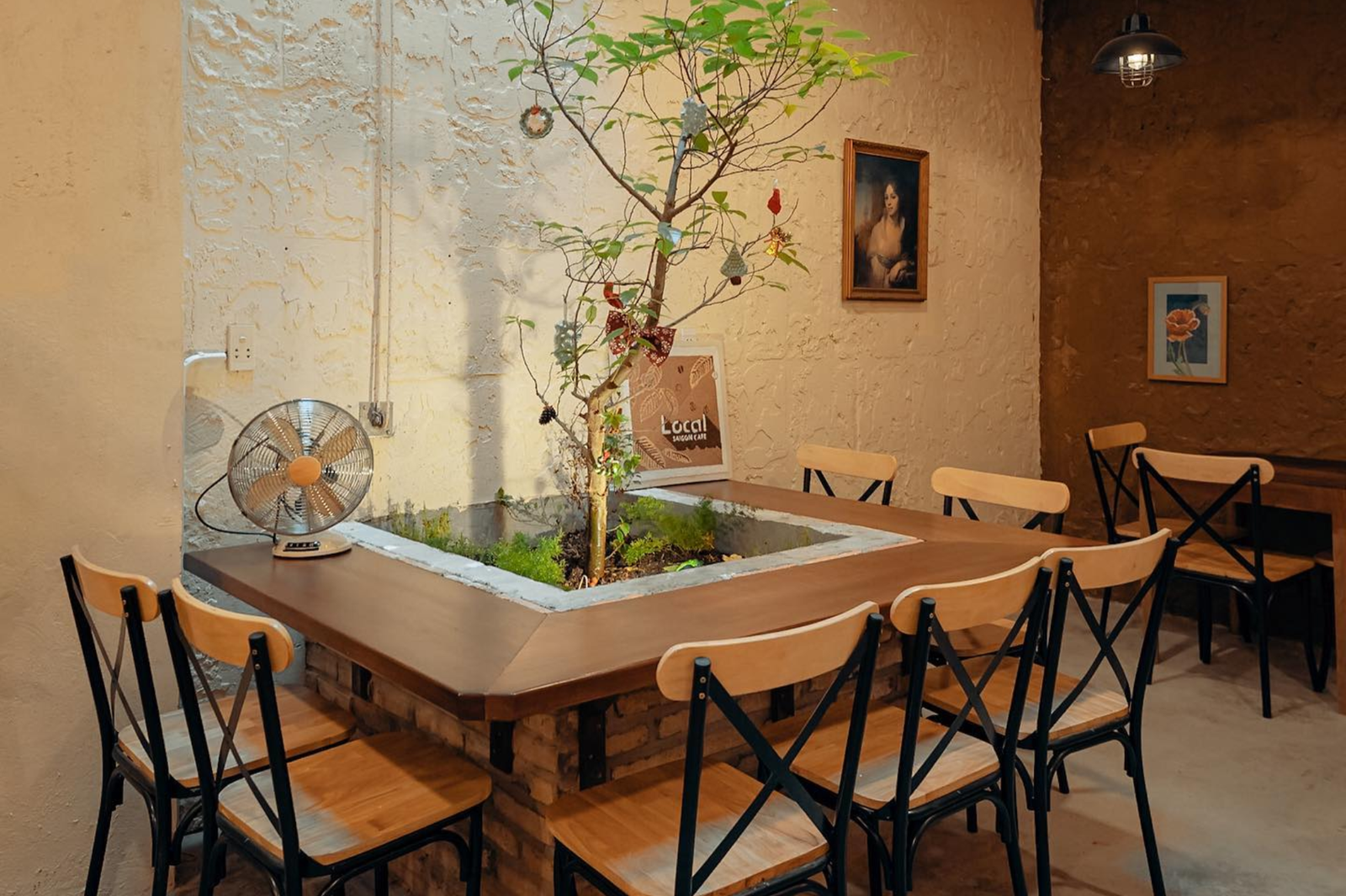 Quán cà phê với bàn ghế gỗ, cây xanh trang trí giữa bàn, tranh treo tường và quạt nhỏ đặt trên bàn.