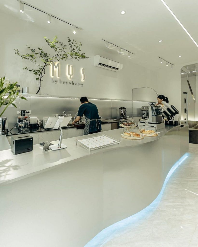 Quán cà phê hiện đại với quầy bar trắng, nhân viên đang pha chế, cây xanh trang trí trên tường.