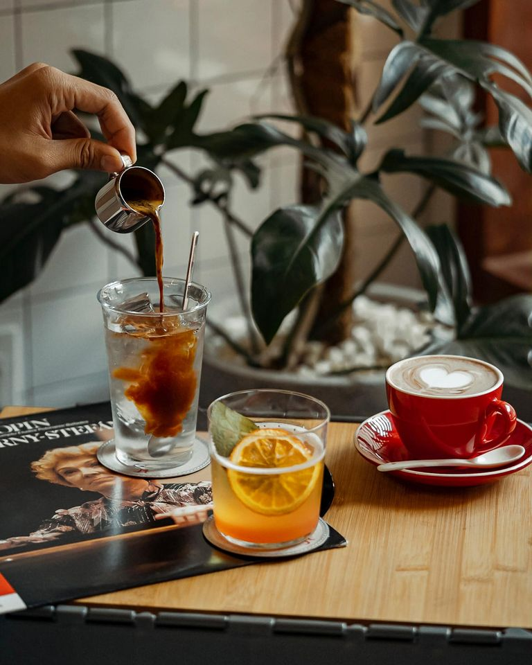 Một tay đang rót cà phê vào ly, bên cạnh là một ly nước cam và một tách cappuccino trên bàn gỗ.