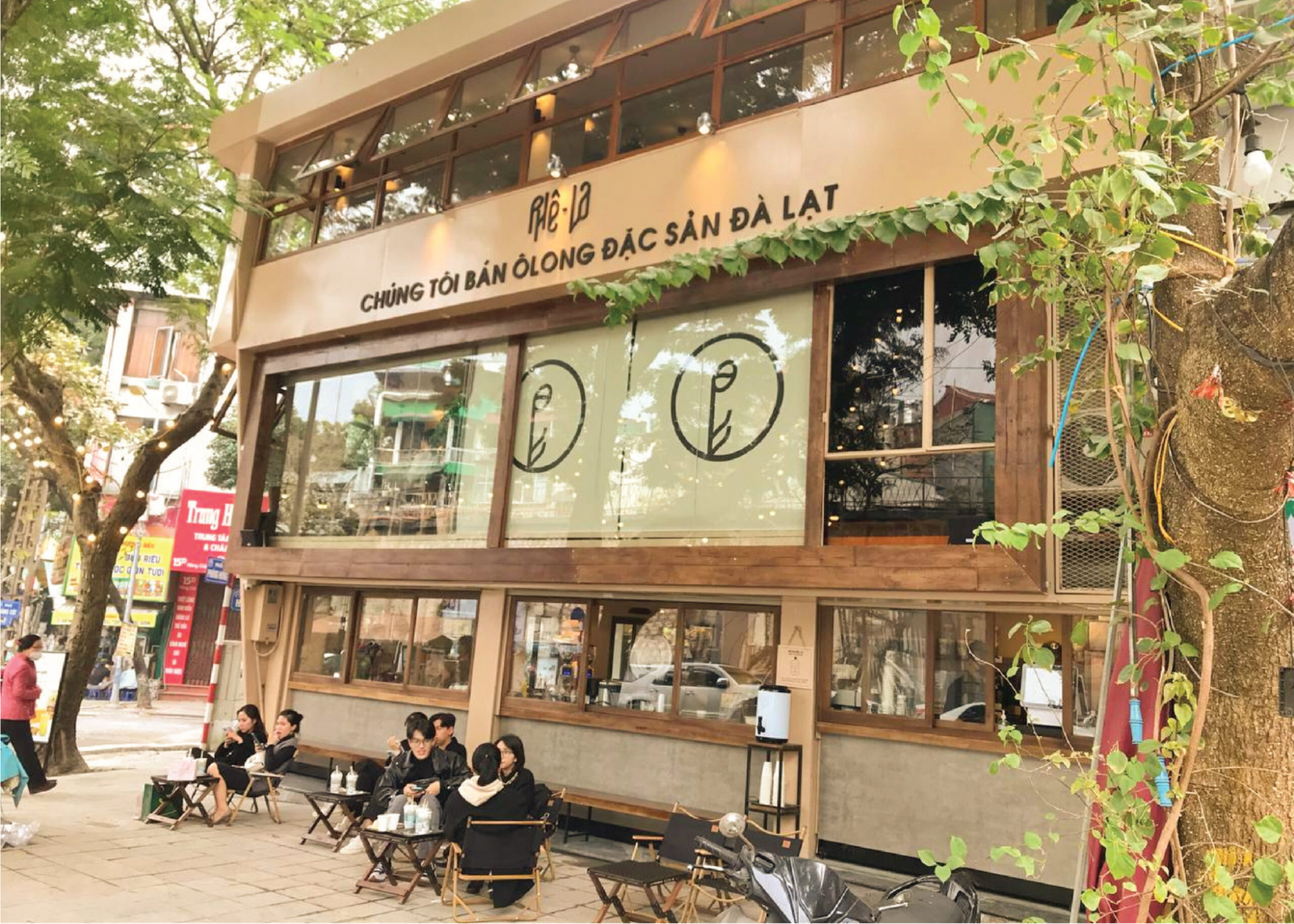 Mặt tiền quán cà phê với khách ngồi ngoài trời, cây xanh xung quanh, tòa nhà hai tầng, biển hiệu lớn.