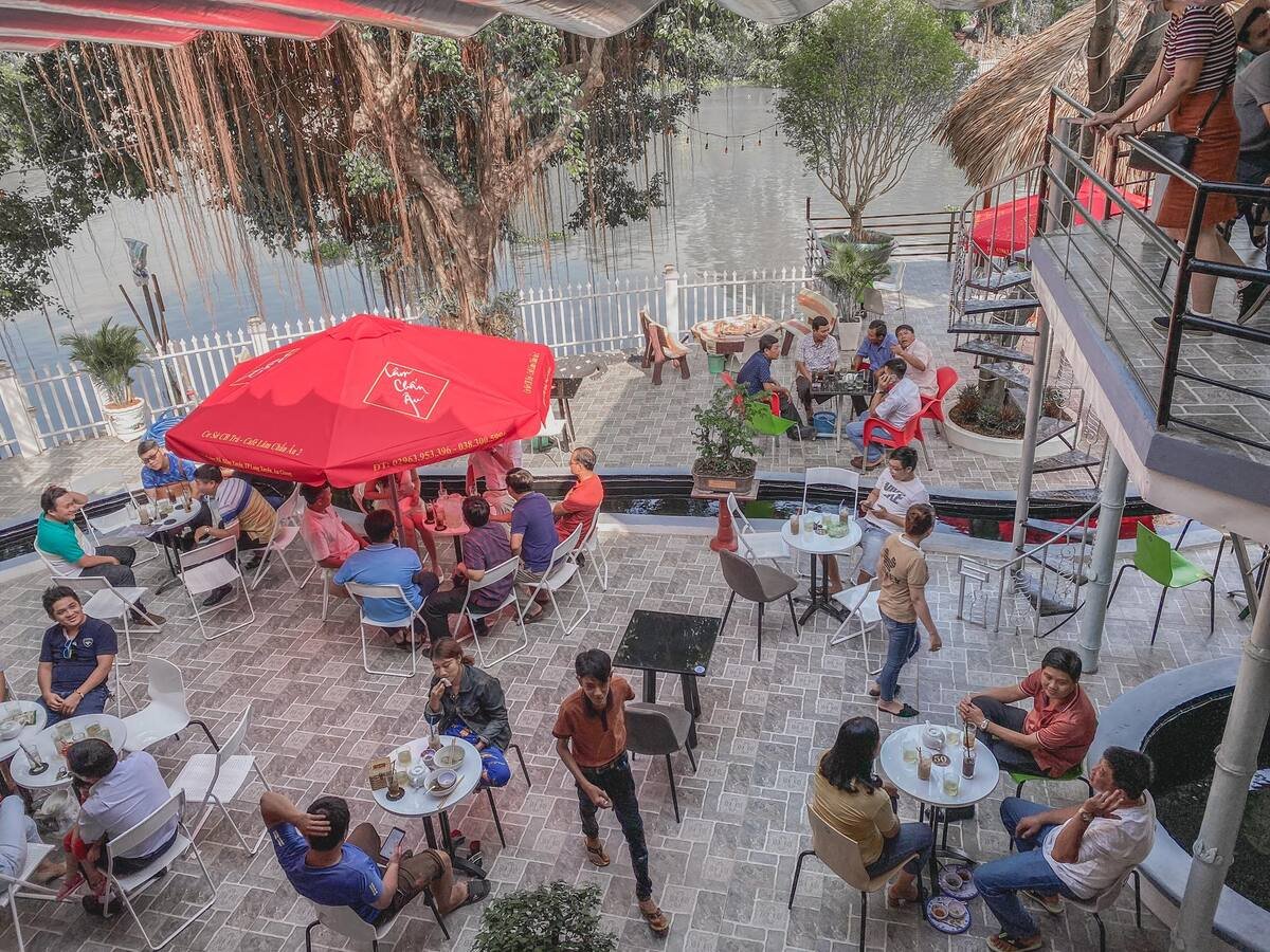 Qu&aacute;n c&agrave; ph&ecirc; ngo&agrave;i trời, kh&aacute;ch ngồi dưới d&ugrave; đỏ, xung quanh l&agrave; c&acirc;y cối v&agrave; hồ nước, kh&ocirc;ng kh&iacute; nhộn nhịp.