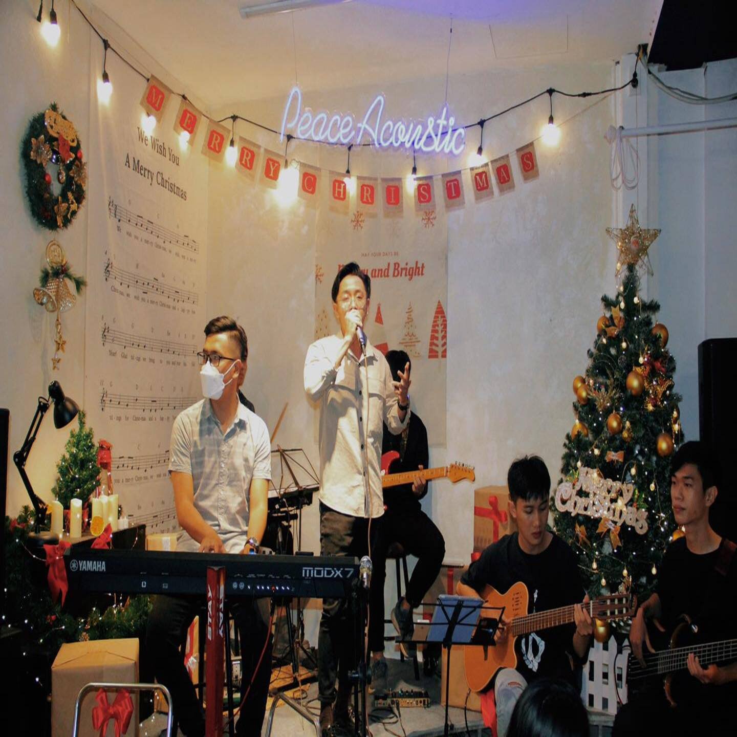 Nh&oacute;m nhạc biểu diễn trong kh&ocirc;ng gian Gi&aacute;ng sinh với c&acirc;y th&ocirc;ng trang tr&iacute; v&agrave; nhạc cụ, c&oacute; người đeo khẩu trang.