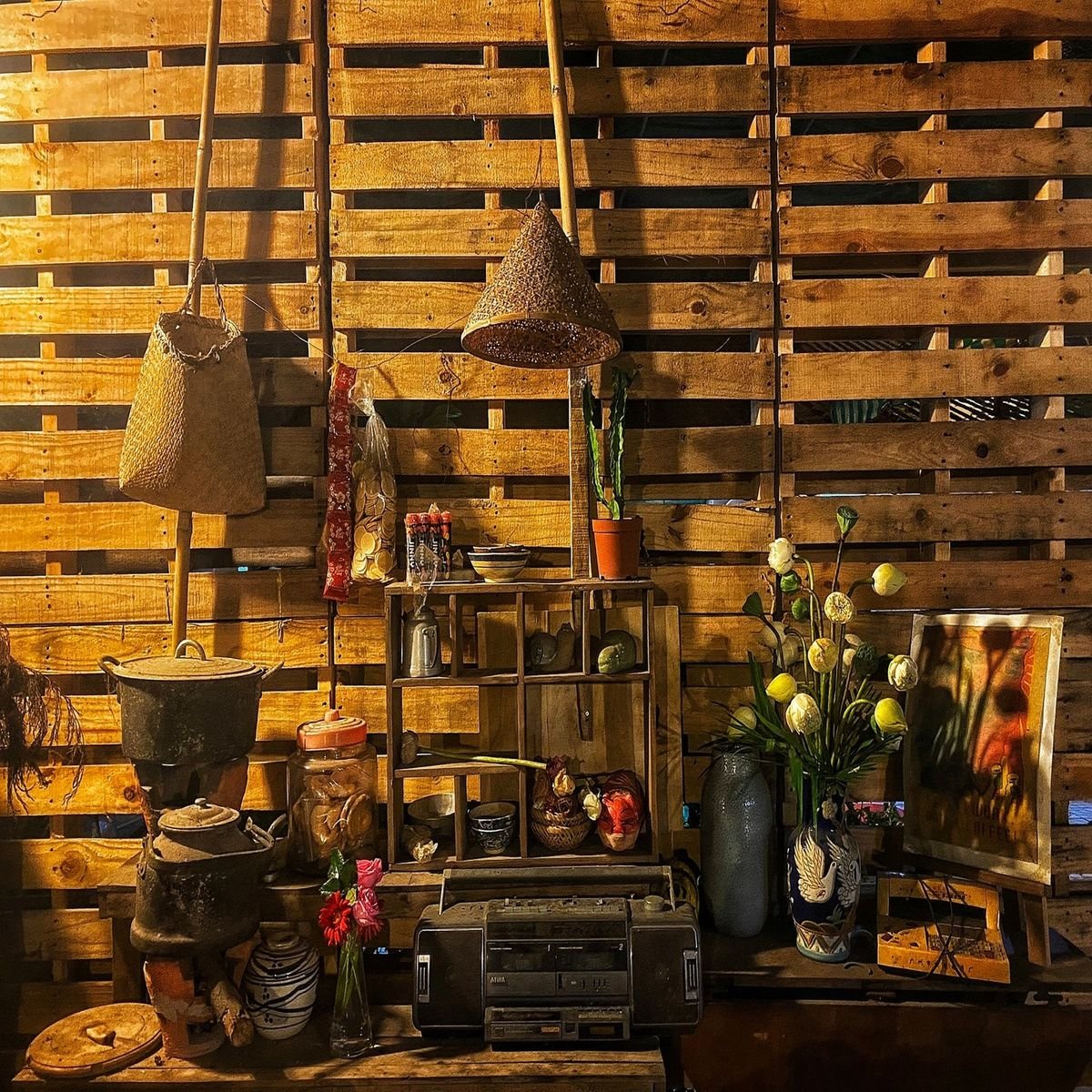 Gian bếp truyền thống với nồi đất, giỏ treo, kệ gỗ chứa đồ, đ&egrave;n lồng tre, b&igrave;nh hoa v&agrave; m&aacute;y cassette cũ.