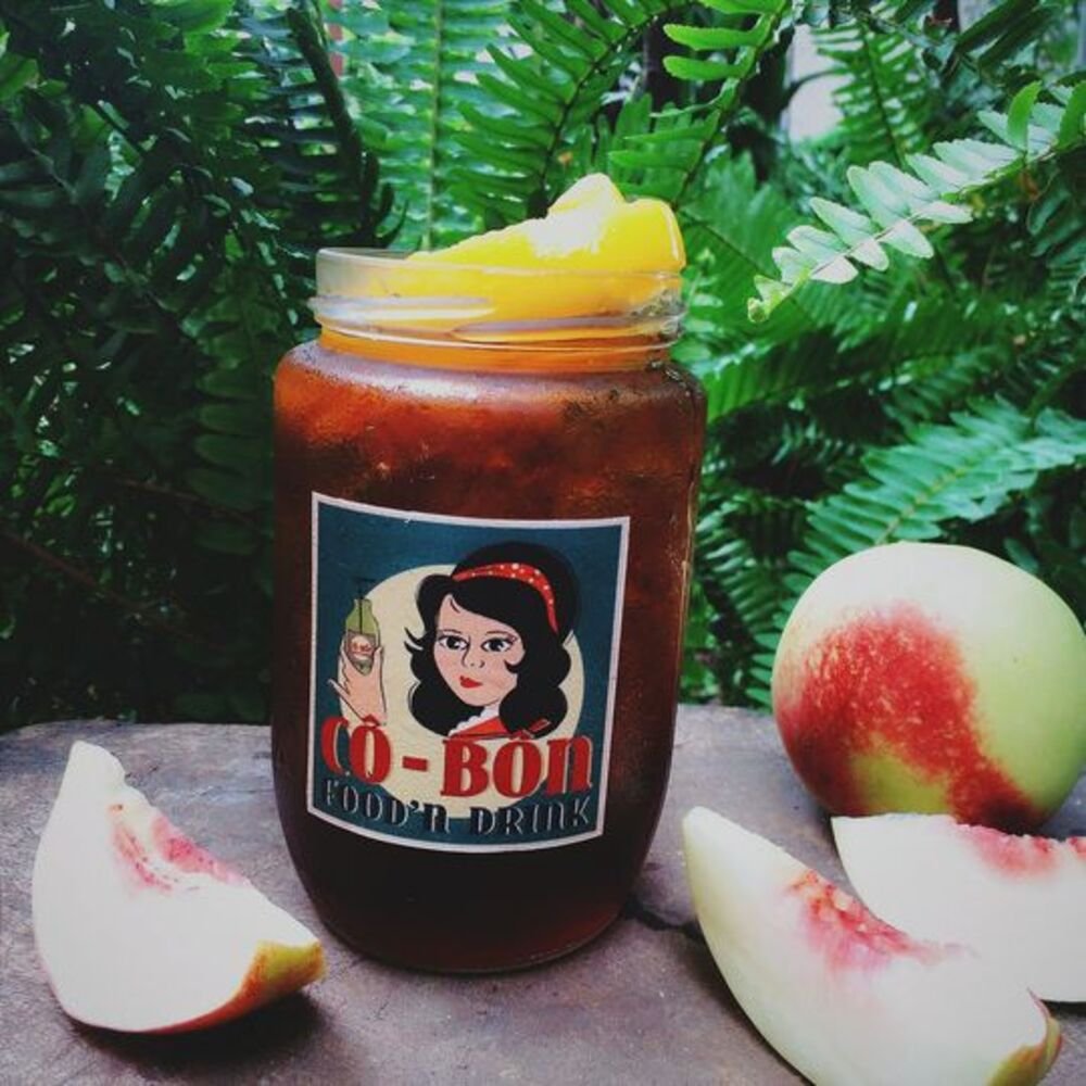 Lọ nước giải kh&aacute;t với l&aacute;t đ&agrave;o tươi tr&ecirc;n miệng, b&ecirc;n cạnh l&agrave; miếng đ&agrave;o, nền l&agrave; c&acirc;y xanh.