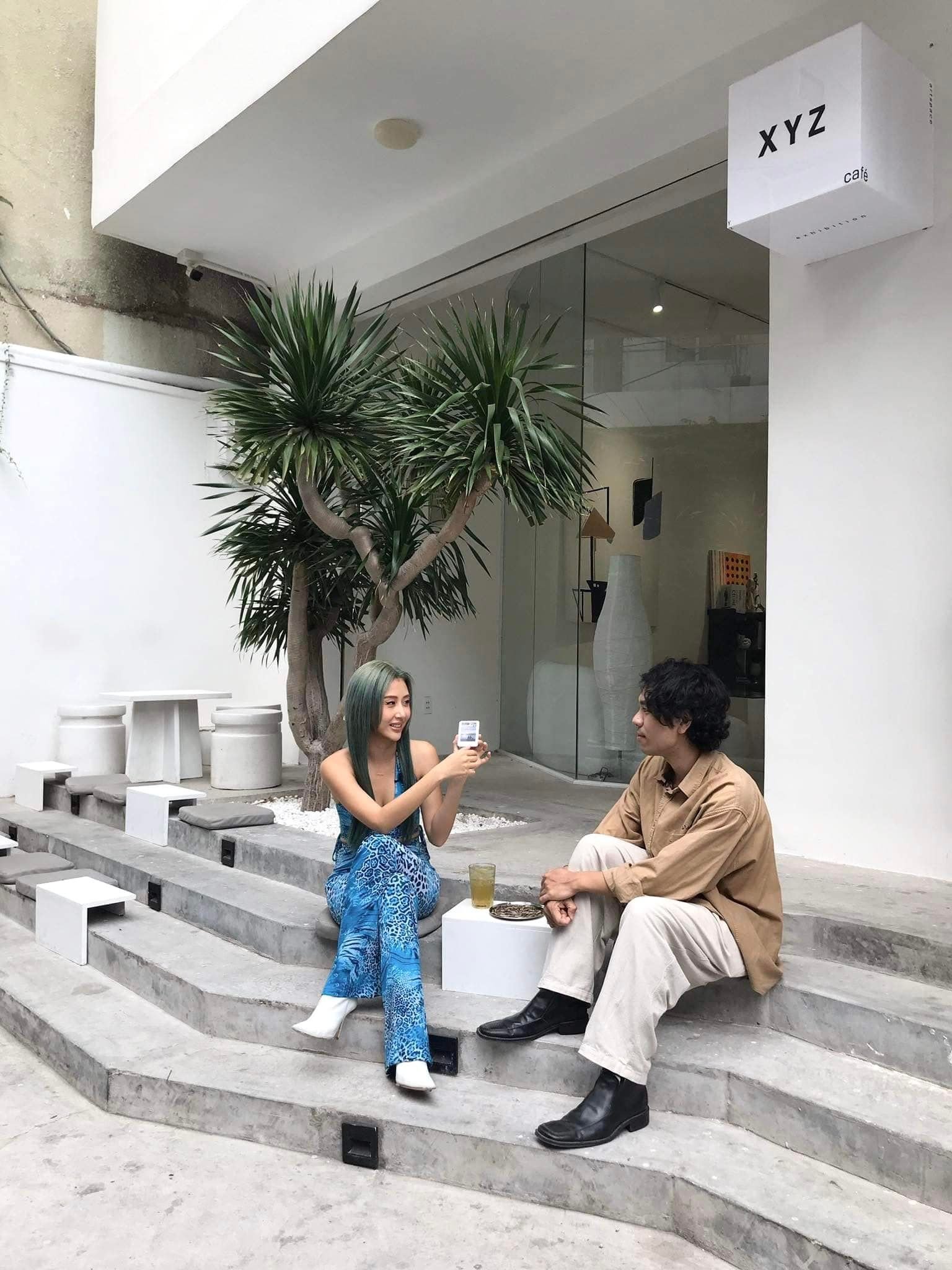 Hai người ngồi trò chuyện trước quán cà phê, cạnh cây xanh, một người cầm điện thoại.