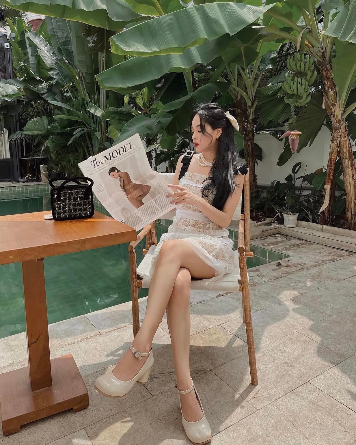 Người phụ nữ ngồi cạnh hồ bơi, đọc tạp chí. Xung quanh là cây chuối xanh tươi và bàn gỗ.