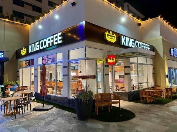 Top 32 thương hiệu nhượng quyền cafe siêu lợi nhuận 22 Quán cà phê với bảng hiệu sáng đèn vào ban đêm, bàn ghế ngoài trời và đèn trang trí quanh mái hiên.