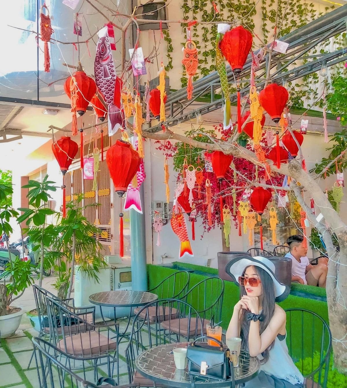 Qu&aacute;n c&agrave; ph&ecirc; trang tr&iacute; đ&egrave;n lồng đỏ v&agrave; c&aacute; giấy, c&oacute; người ngồi b&agrave;n ngo&agrave;i trời, c&acirc;y xanh xung quanh.