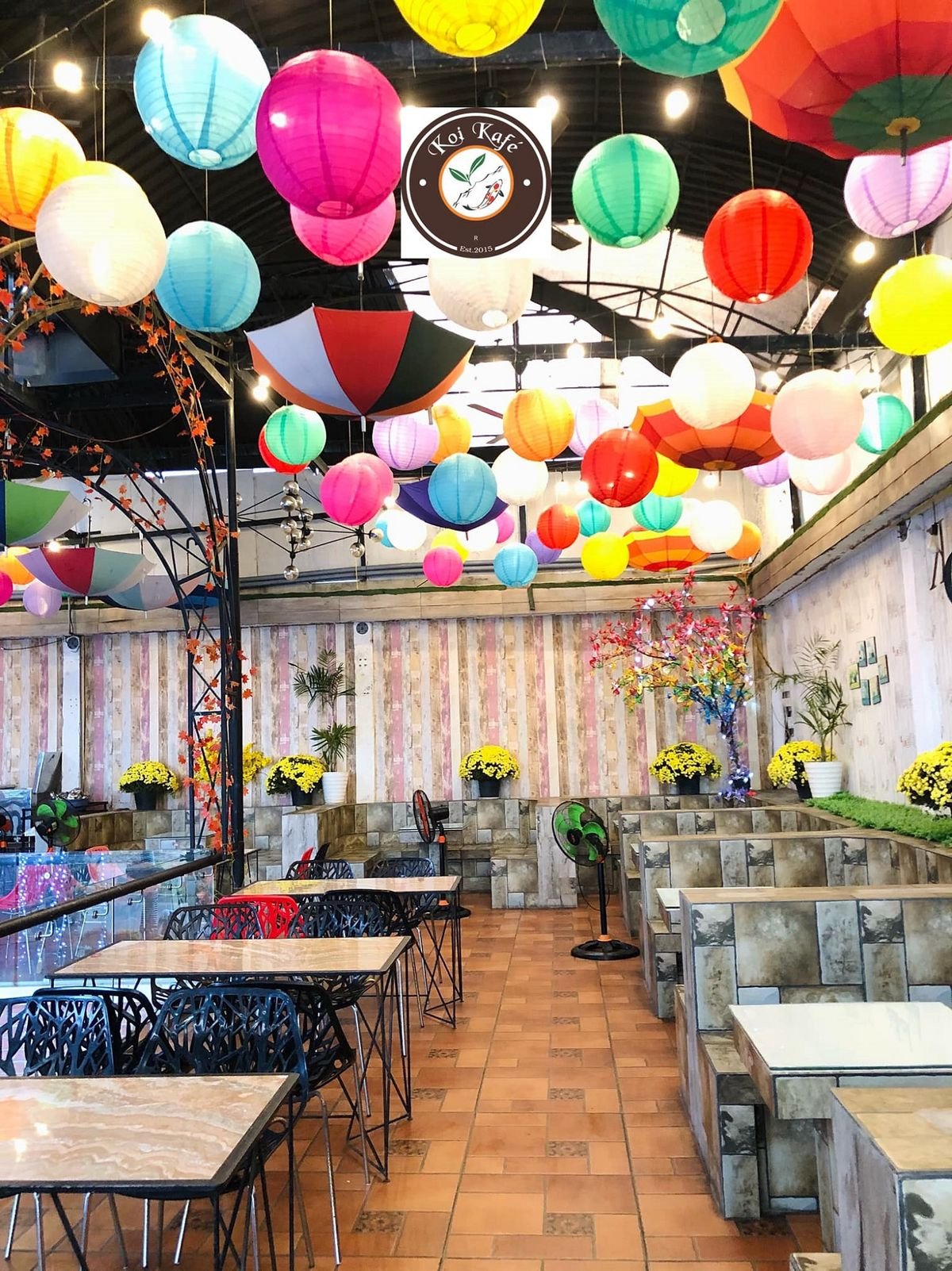 Qu&aacute;n c&agrave; ph&ecirc; với trần nh&agrave; treo nhiều lồng đ&egrave;n v&agrave; &ocirc; m&agrave;u sắc rực rỡ, b&agrave;n ghế sắp xếp gọn g&agrave;ng.