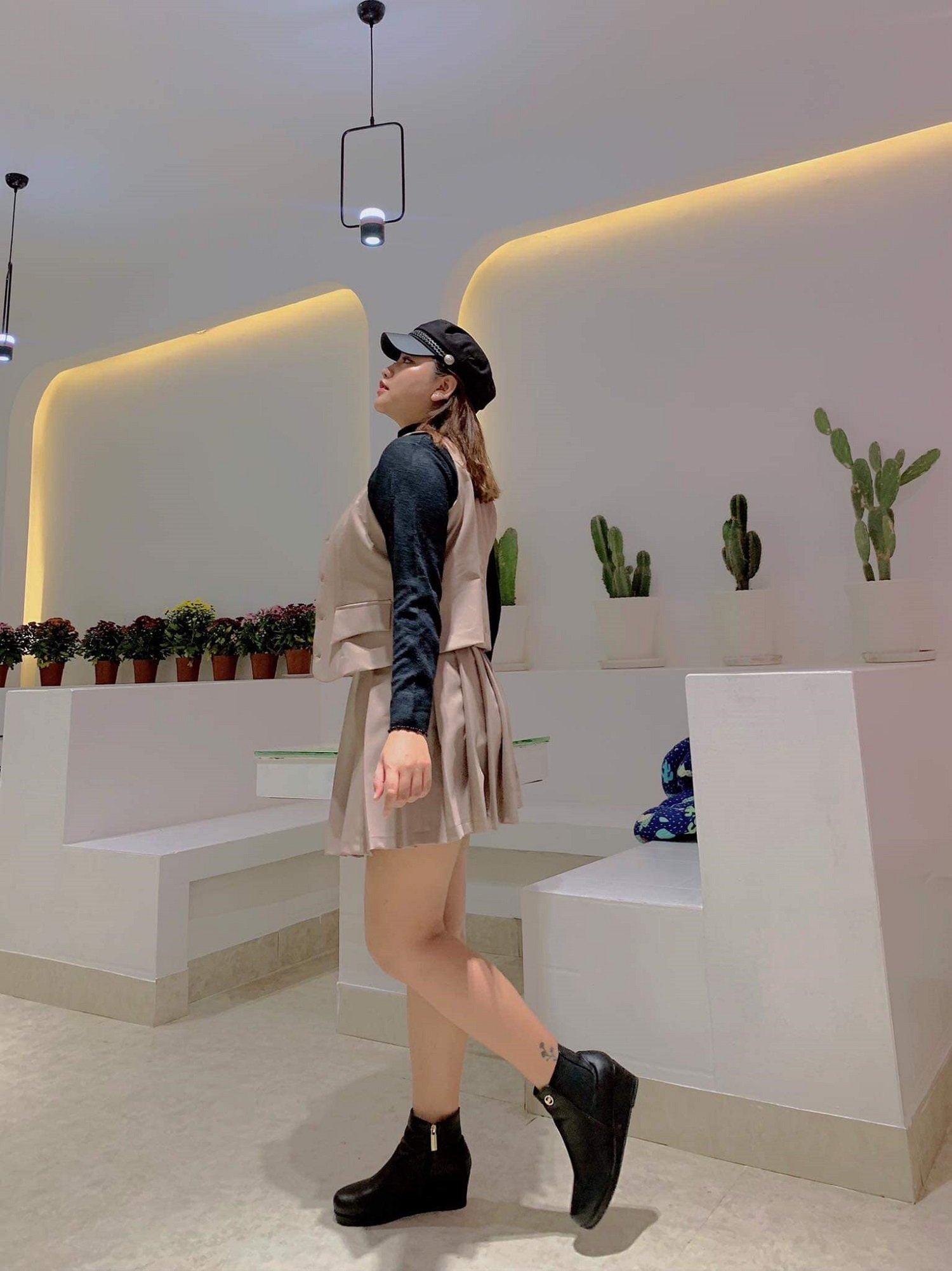 Người phụ nữ mặc v&aacute;y v&agrave; đội mũ đen đứng trong kh&ocirc;ng gian c&oacute; c&acirc;y xương rồng trang tr&iacute; tr&ecirc;n kệ.