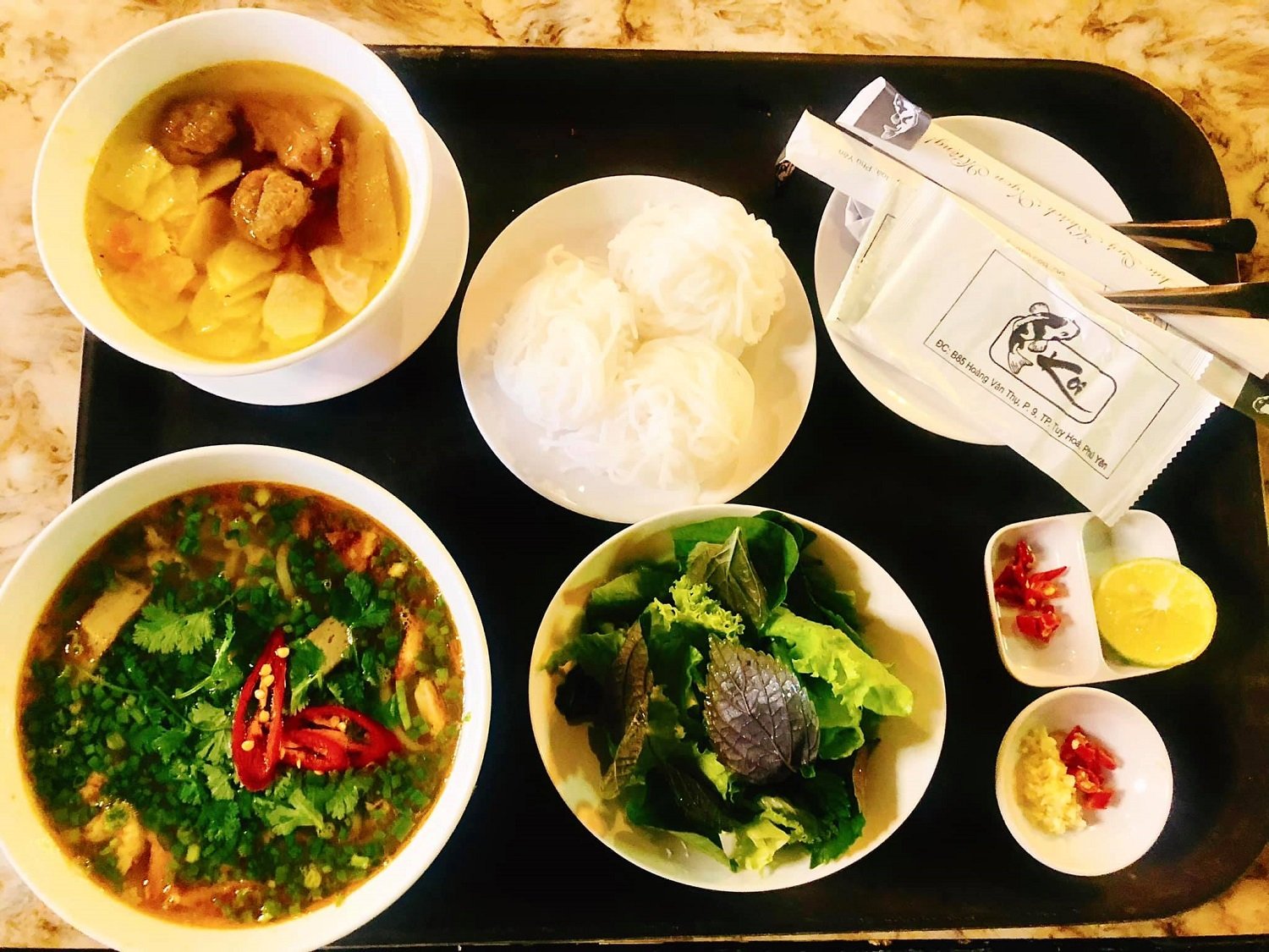 Khẩu phần ăn gồm b&uacute;n, canh thịt, rau sống, nước chấm v&agrave; gia vị tr&ecirc;n khay, với đũa v&agrave; khăn giấy b&ecirc;n cạnh.