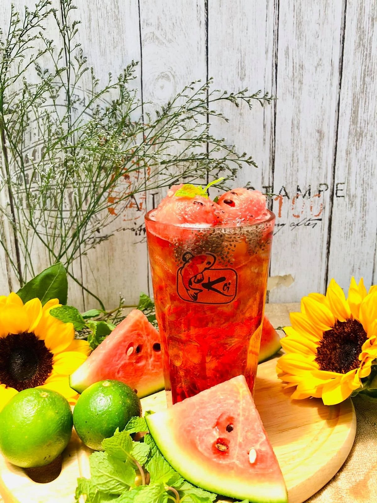 Ly tr&agrave; tr&aacute;i c&acirc;y với l&aacute;t dưa hấu, chanh v&agrave; bạc h&agrave;, b&ecirc;n cạnh hoa hướng dương tr&ecirc;n nền gỗ trắng.